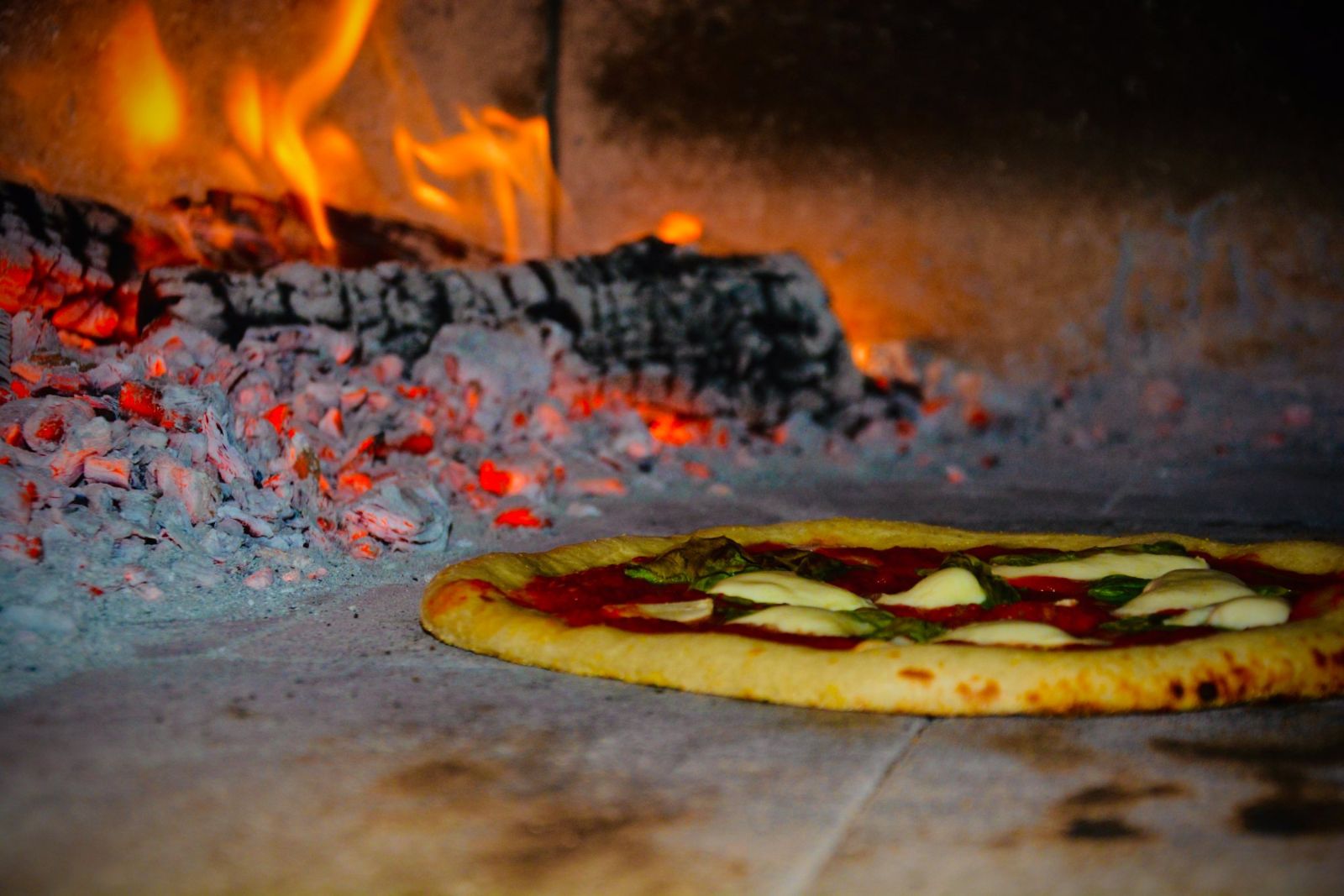 A margherita pizza baking next to glowing embers in the wood-fired oven