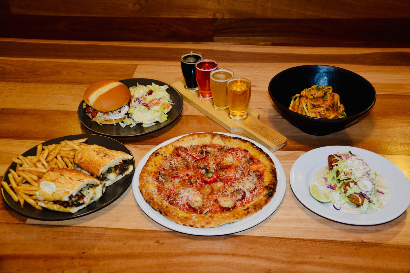 A table full of cheesesteak, pizza, burger, rockfish taco, pasta and a beer flight